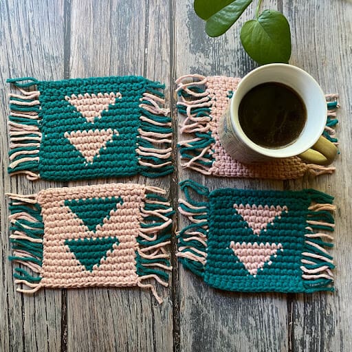 Overhead image of 4 crochet coasters with a triangular geometric design and fringe on 2 opposite sides. Two are in peach with teal triangles and two are in teal with peach triangles - a mug sits on one.