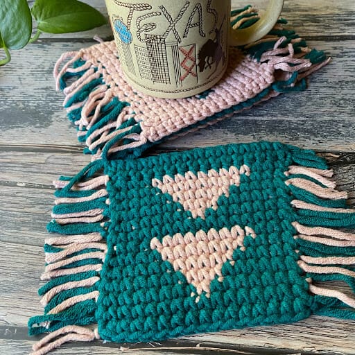 Image of 2 crochet coasters with a triangular geometric design and fringe on 2 opposite sides. One is in peach with teal triangles and the other is in teal with peach triangles - a mug sits on one and a plant is in the background.