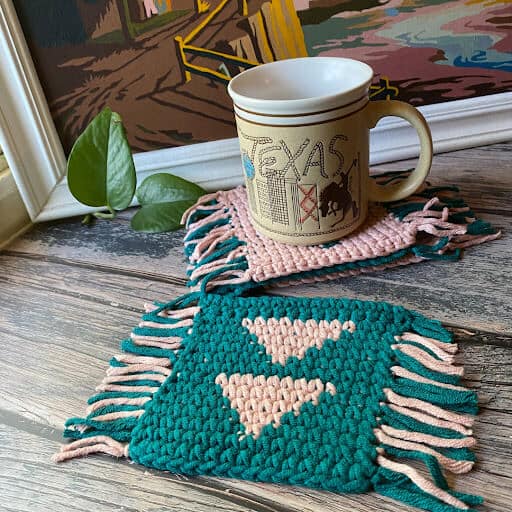 Image of 2 crochet coasters with a triangular geometric design and fringe on 2 opposite sides. One is in peach with teal triangles and the other is in teal with peach triangles - a mug sits on one and a painting is in the background.